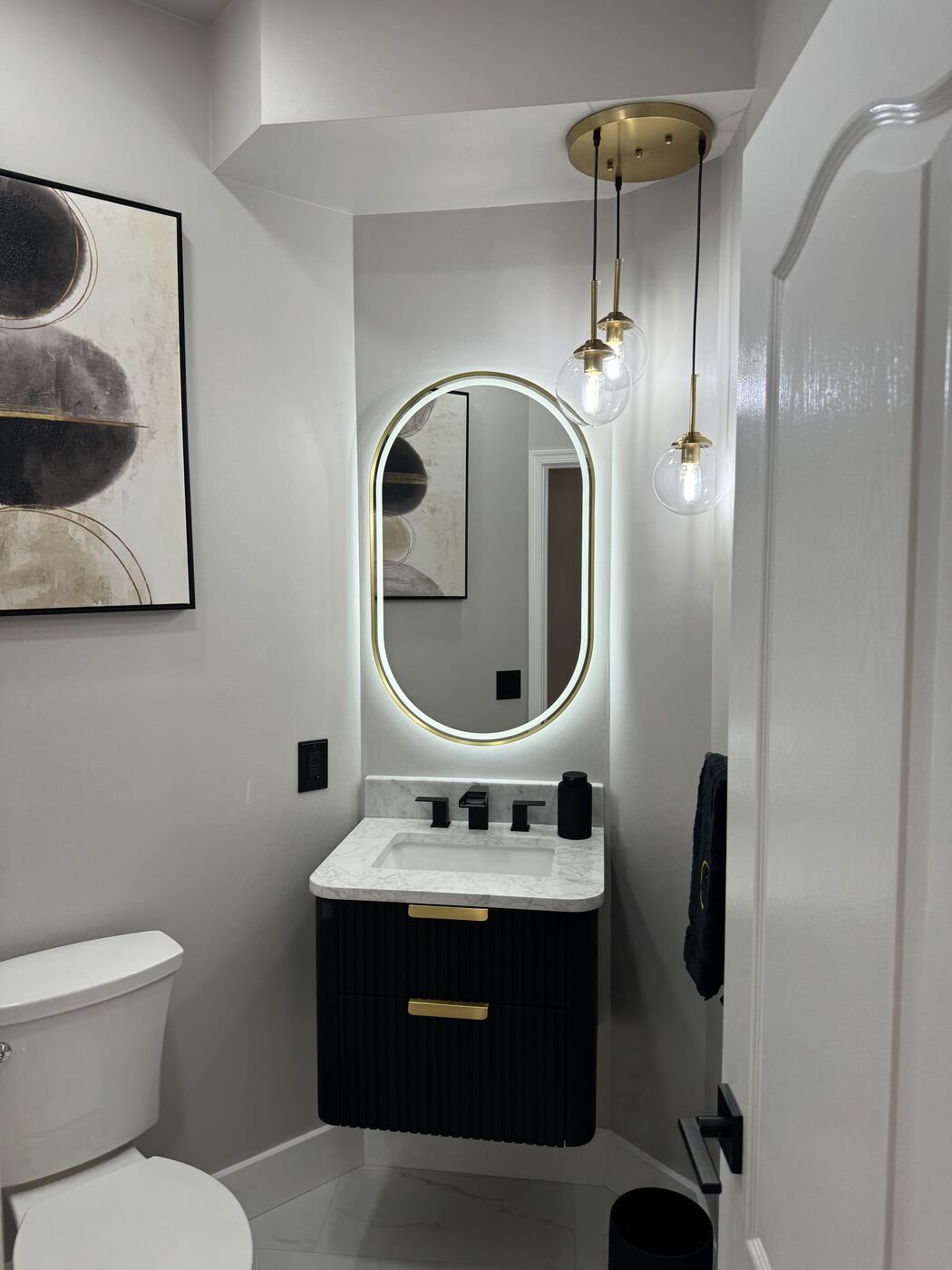 Powder room with black fluted vanity, backlit oval mirror and brass pendant lights